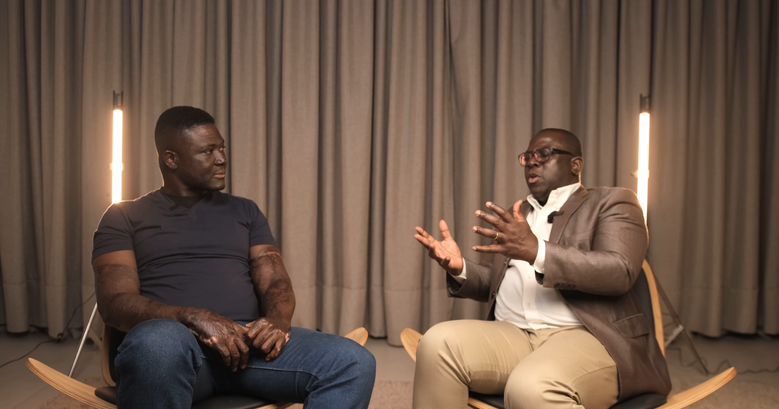 Pierre is seated with Naa Oyoo Kortey of Channel1 in a studio set having a conversation, with a large screen showing a smiling man in the background.