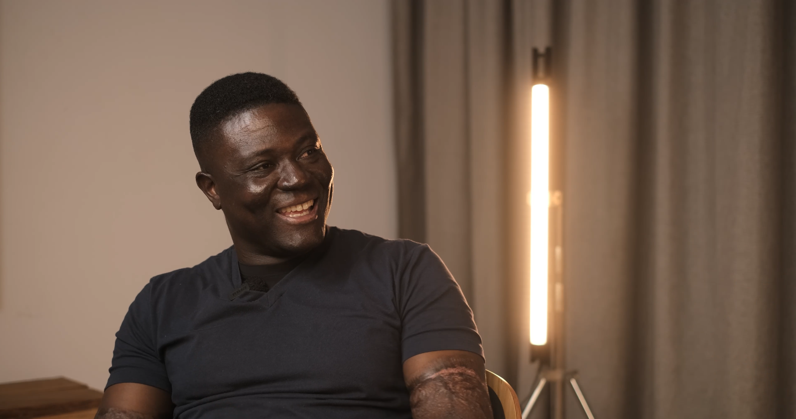 Pierre is seated with LexisBill of JoyFM in purple chairs having a conversation in a radio studio with a large screen showing a promotional image for a personality profile show.