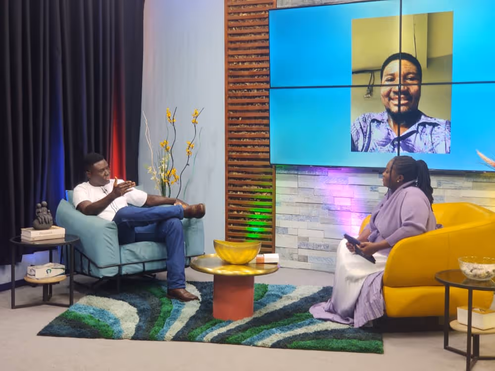 Pierre is seated with Naa Oyoo Kortey of Channel1 in a studio set having a conversation, with a large screen showing a smiling man in the background.