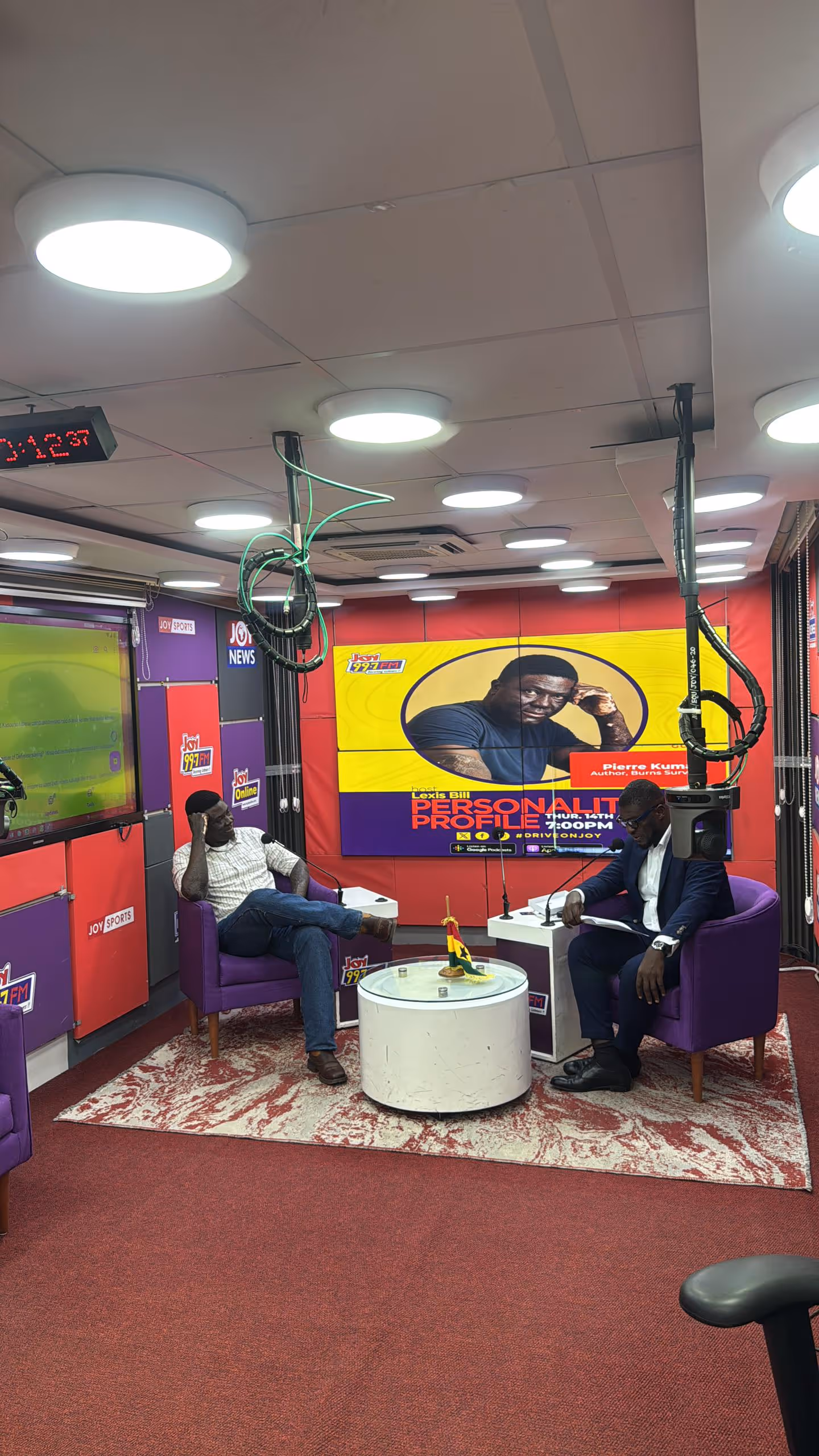 Pierre is seated with LexisBill of JoyFM in purple chairs having a conversation in a radio studio with a large screen showing a promotional image for a personality profile show.